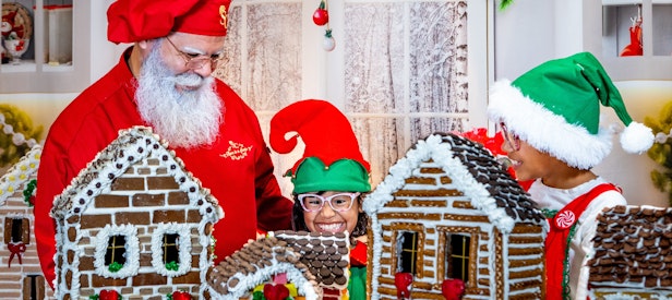 two children dressed as elves join Santa as they make gingerbread houses